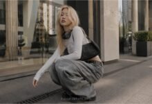 Acubi baggy jeans outfit with grey loose denim, fitted white long sleeve top, black shoulder bag, headphones, and chunky shoes in a modern city setting