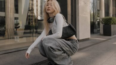 Acubi baggy jeans outfit with grey loose denim, fitted white long sleeve top, black shoulder bag, headphones, and chunky shoes in a modern city setting