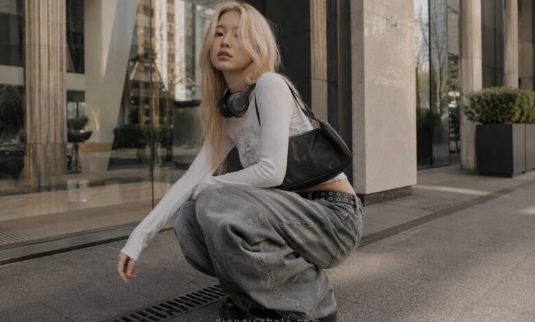Acubi baggy jeans outfit with grey loose denim, fitted white long sleeve top, black shoulder bag, headphones, and chunky shoes in a modern city setting