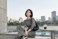 Acubi color palette outfit with grey fitted top, muted green denim, black shoulder bag, black sneakers, silver jewelry, and modern city terrace styling