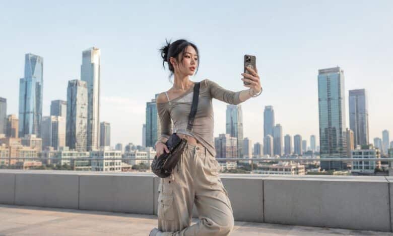 Woman taking a selfie in an Acubi outfit with asymmetrical grey top, beige cargo pants, crossbody bag, silver jewelry, and chunky sneakers