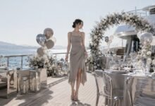 Acubi wedding guest on a private yacht wearing a grey asymmetrical dress with silver jewelry, metallic mini bag, sleek heels, chrome decor, floral arch, and silver balloons