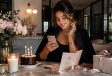 Stylish woman reading a birthday message on her phone beside cake, candles, flowers, gift box, and a birthday card