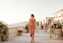 Woman wearing a coral satin destination wedding guest dress on an elegant seaside terrace at sunset