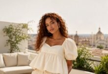 Woman with curly red hair wearing a cream babydoll top in a sunlit rooftop fashion setting