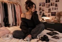 Fashion blogger sitting in a cozy bedroom closet with moodboard wall, clothing rack, oversized sweater, loose jeans, sneakers, headphones, camera, and accessories