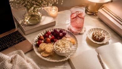 Aesthetic desk with pretty snacks, strawberries, grapes, crackers, cheese, popcorn, pink drink, notebook, laptop, candle, and warm study setup
