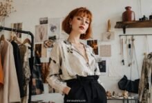 Tiny Brands With Main-Character Energy 3 Stylish young woman with red bob haircut in an indie fashion atelier, wearing unique printed blouse and tailored black trousers among handmade clothing and moodboard details