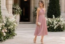 Stylish wedding guest in a soft pink midi dress outside an elegant garden wedding venue with florals and stone architecture