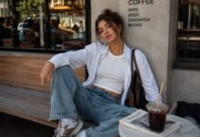 Stylish young woman in a white fitted tee, relaxed striped shirt, loose denim, sneakers, and brown shoulder bag at a cozy urban café.