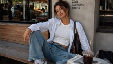 Stylish young woman in a white fitted tee, relaxed striped shirt, loose denim, sneakers, and brown shoulder bag at a cozy urban café.