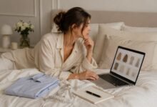 Stylish woman shopping for clothes online from bed while comparing fashion items, measurements and outfit notes